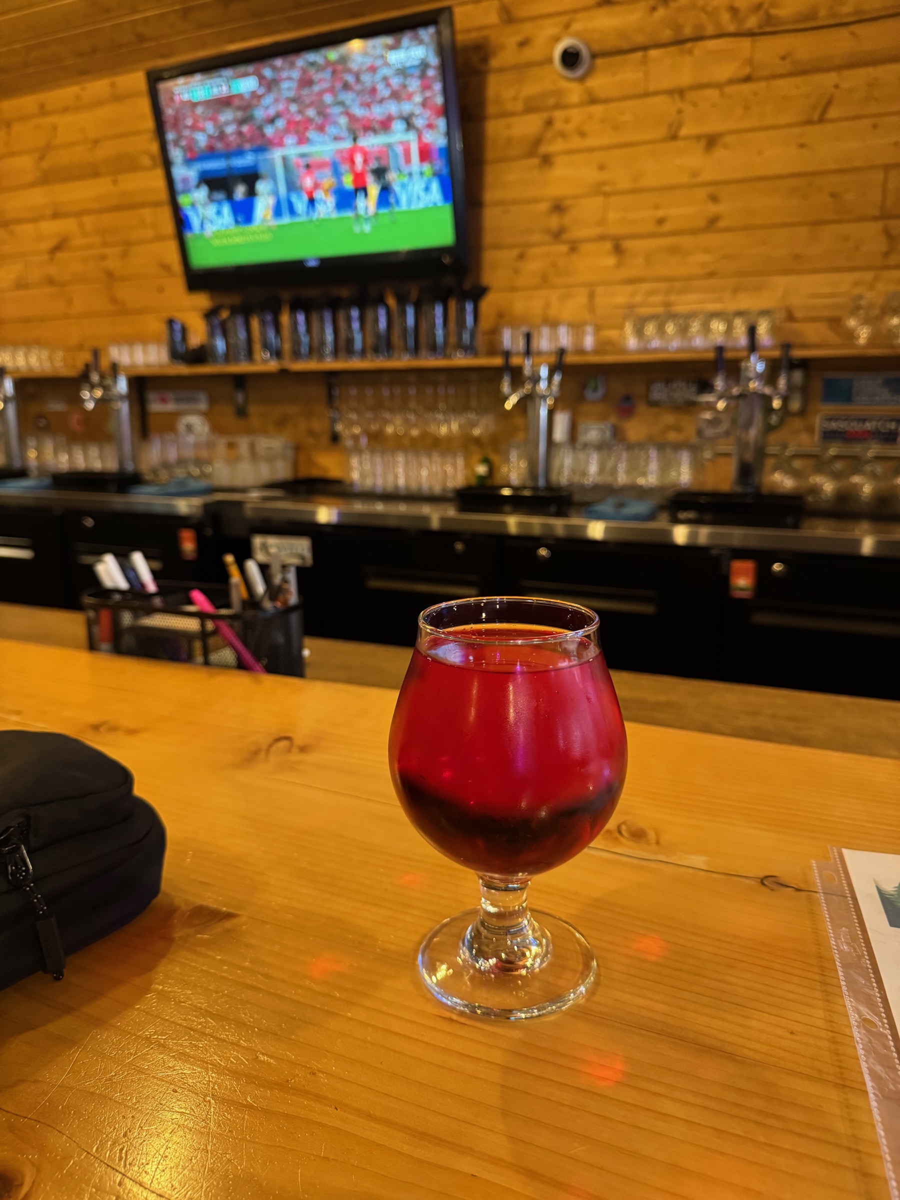A glass of amber-red cider glowing on the wood bar of the West End Tap Room in Forks, with a football match on the screen behind